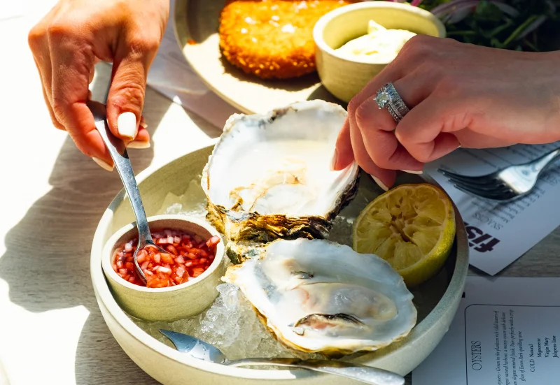 Person eating fresh oysters with lemon and red sauce at an outdoor meal, with bread and butter visible in the background