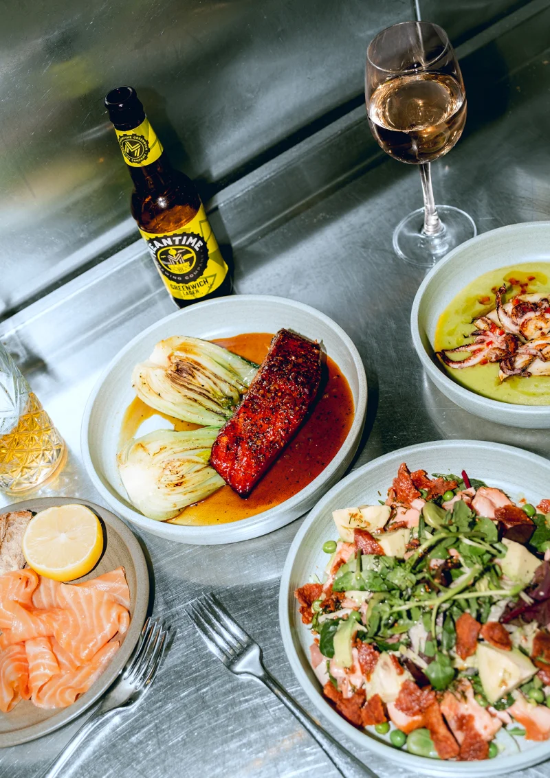 Overhead view of a seafood meal with salmon, roasted vegetables, and salads served on white plates alongside a beer bottle and glass of white wine on a grey wooden table.